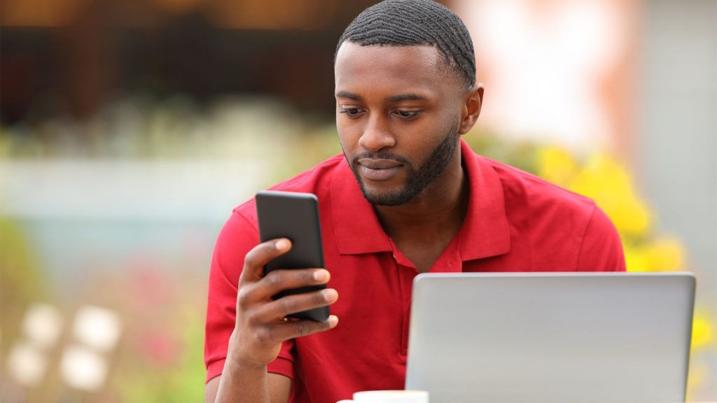 Homem de camisa vermelha olhando atentamente para o celular, com o laptop aberto à sua frente, do lado de fora.