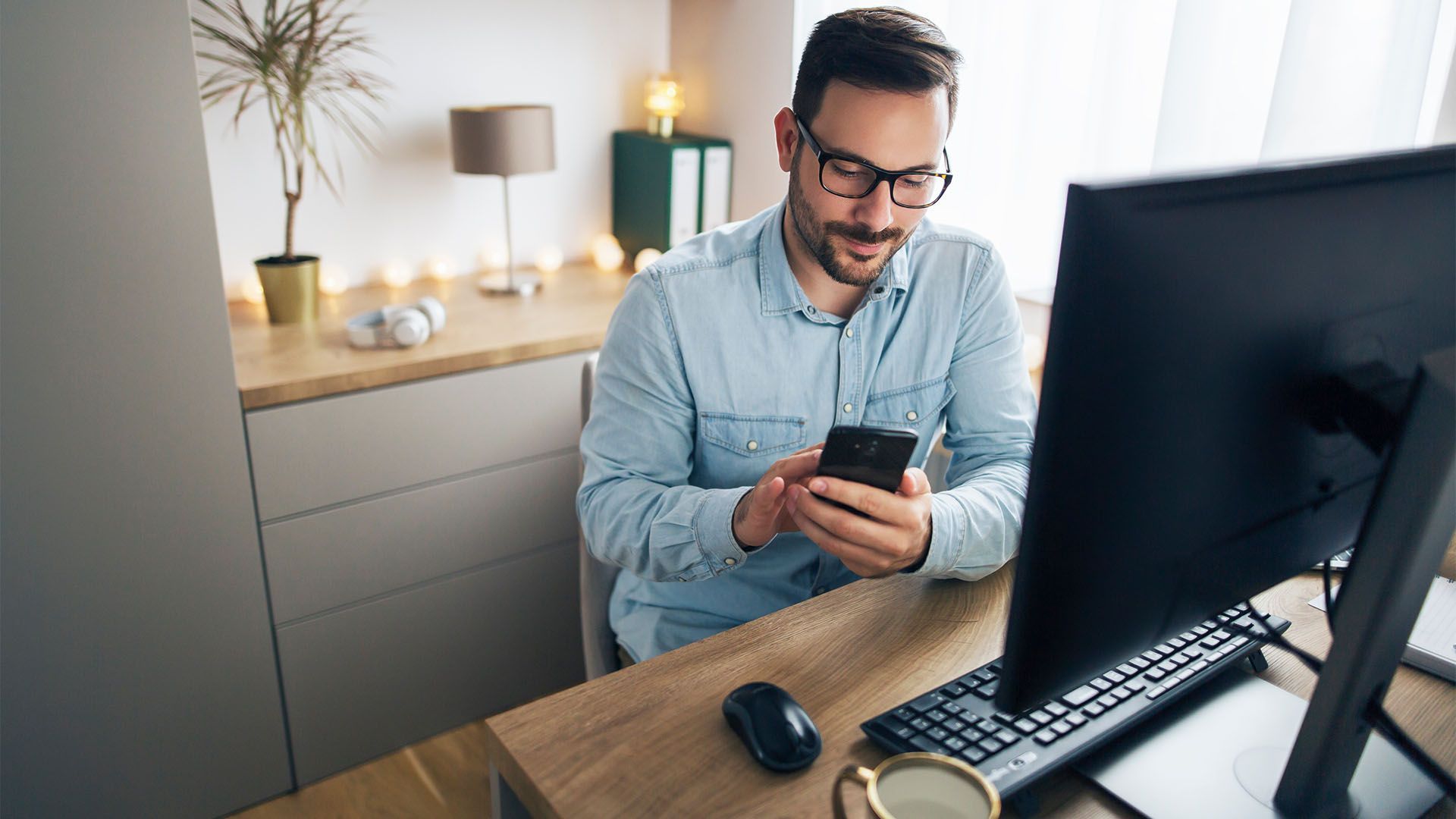 Homem de óculos sorri para o celular enquanto está sentado em uma mesa com um computador, uma caneca e um mouse.