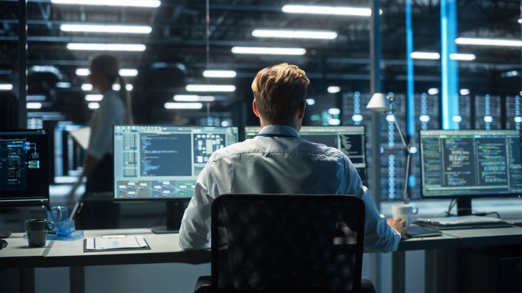 Pessoa trabalhando em um computador com vários monitores em um centro de dados. Servidores e outra pessoa ao fundo.