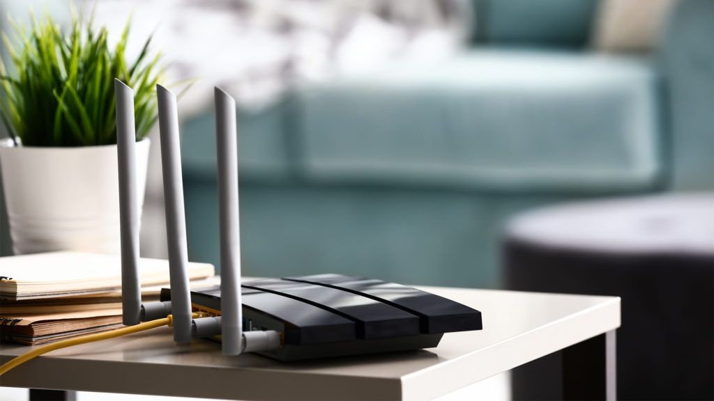 Roteador sem fio preto com três antenas sobre uma mesa branca em uma sala de estar.