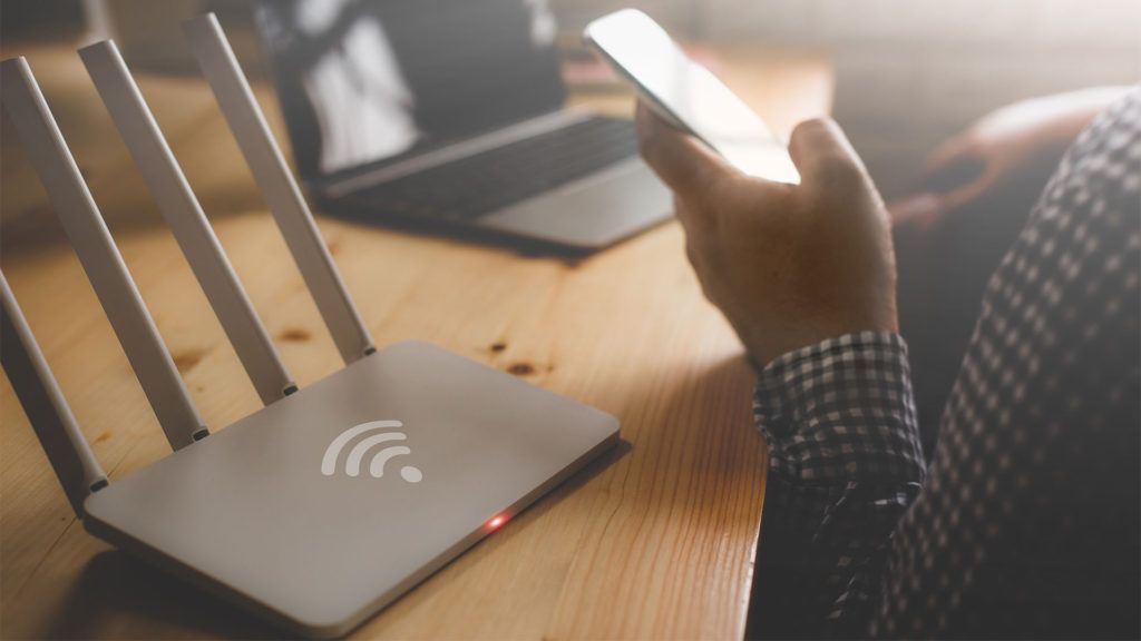 Uma pessoa segurando um celular perto de um roteador Wi-Fi sobre uma mesa de madeira, com um laptop ao fundo.