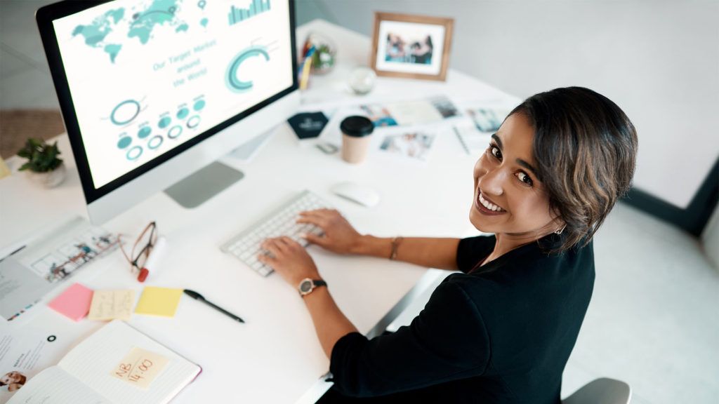 Mulher trabalhando em uma mesa, olhando para cima e sorrindo. A tela do computador exibe gráficos de dados.