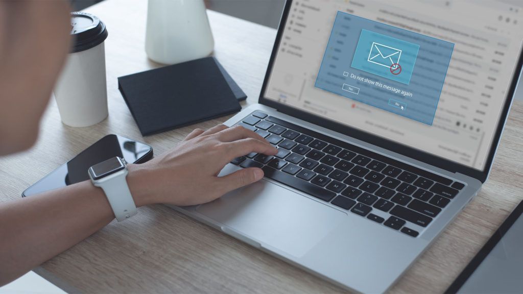 Pessoa digitando em um laptop, redigindo um e-mail, com café, telefone e relógio sobre a mesa.