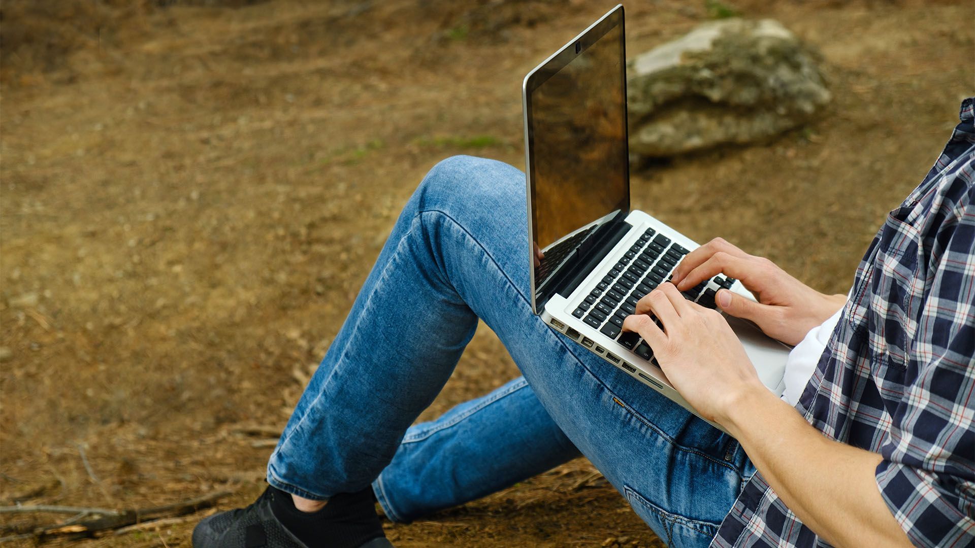 Pessoa sentada ao ar livre, usando um laptop. Vestindo calça jeans e camisa xadrez.