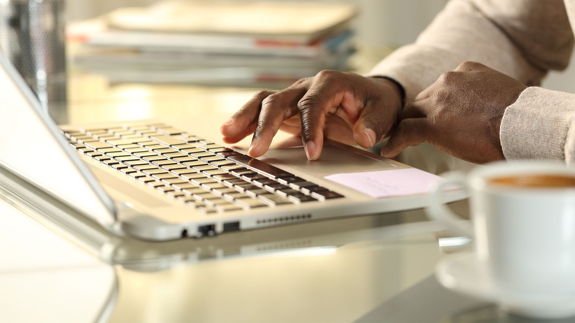 Mãos digitando em um teclado de laptop perto de uma xícara de café sobre uma superfície de vidro.