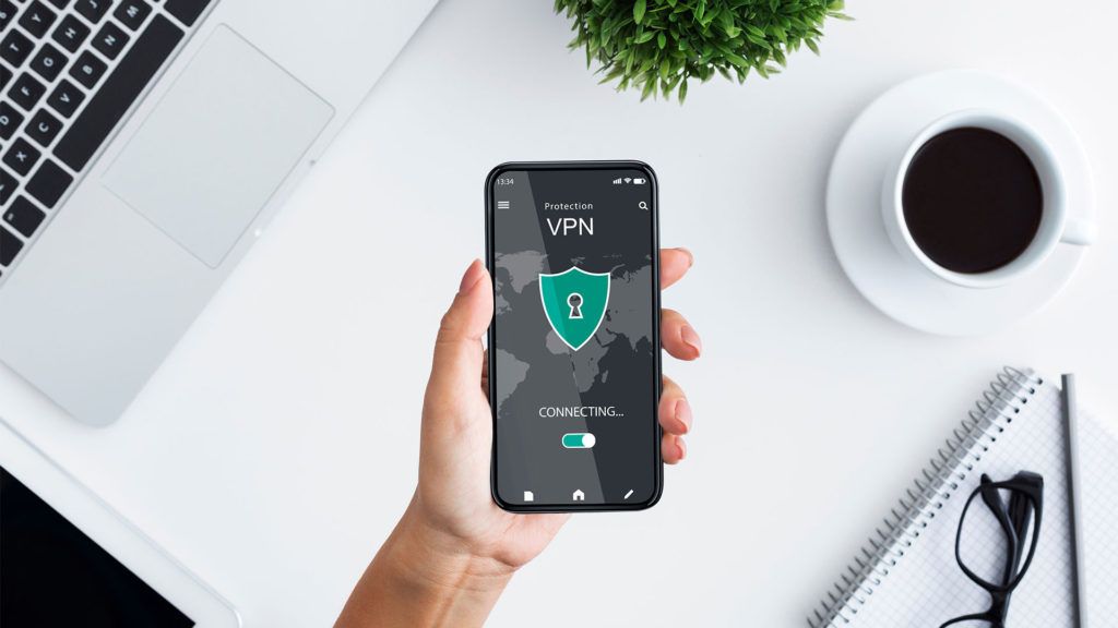 Mão segurando um smartphone exibindo a interface de um aplicativo VPN, sobre uma mesa branca com um laptop, café e uma planta.