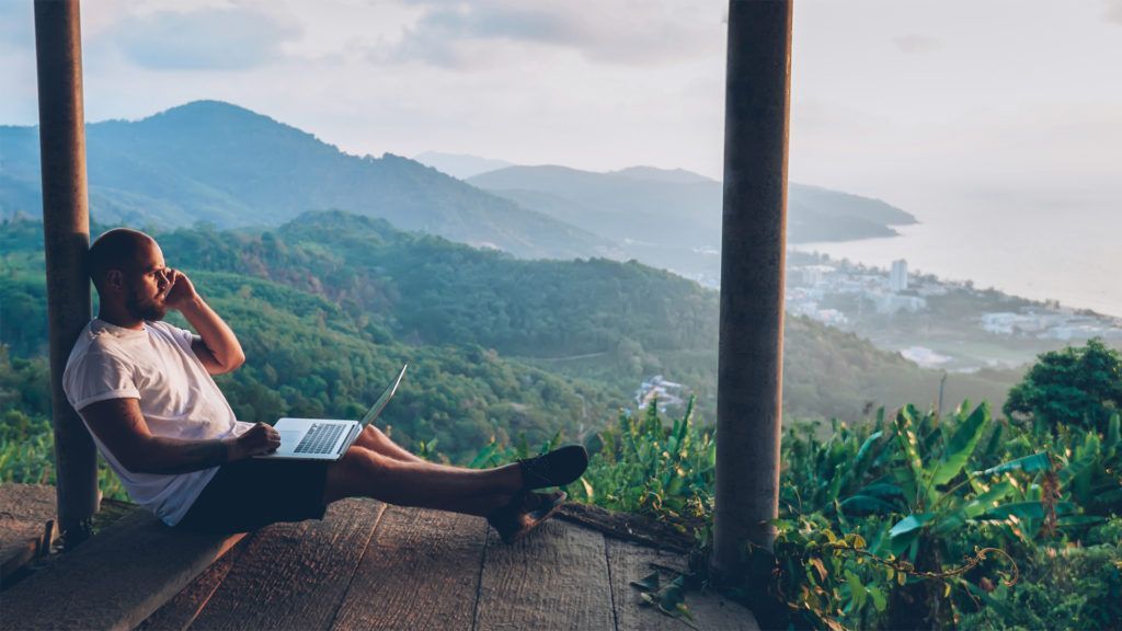 Homem trabalhando em um laptop enquanto fala ao telefone, com vista para a paisagem montanhosa e o oceano.