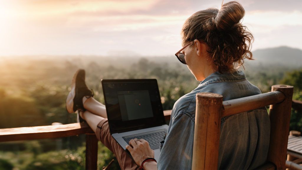 Mulher usando laptop ao ar livre, com os pés apoiados no corrimão da cadeira, com vista para uma paisagem exuberante.