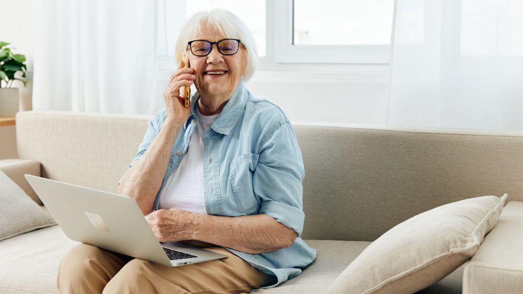 Mulher sentada no sofá, usando um laptop, falando ao telefone e sorrindo. Ambiente interno, bem iluminado.