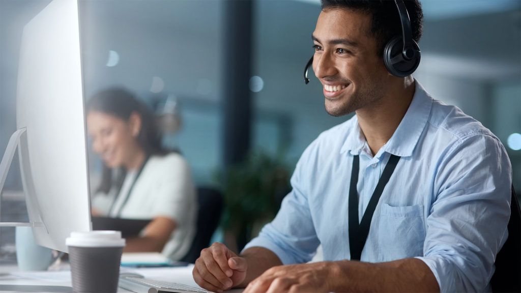 Homem usando fone de ouvido, sorrindo enquanto trabalha no computador em um escritório.