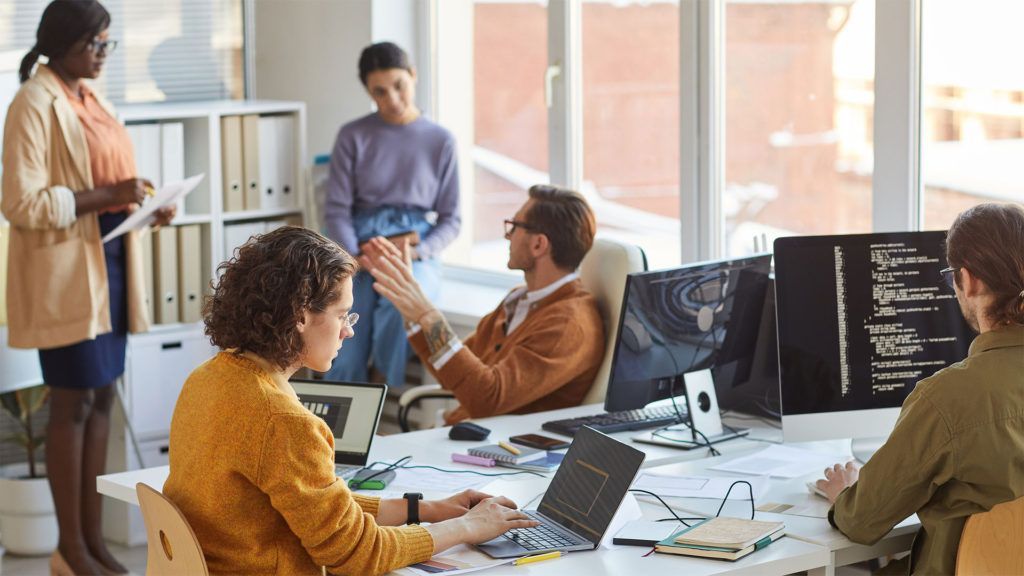 Grupo de pessoas trabalhando em um escritório bem iluminado. Algumas estão em pé, outras sentadas em frente a computadores, colaborando e discutindo ideias.