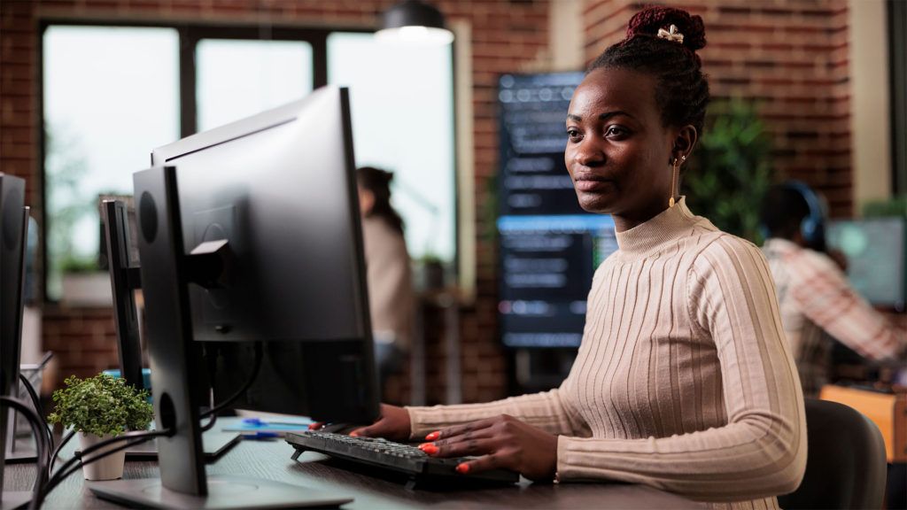 Mulher programando em um computador em um escritório; parede de tijolos ao fundo.