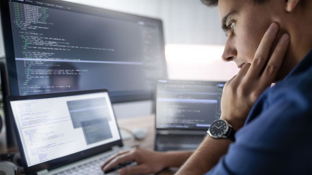 Homem programando em várias telas de computador, expressão concentrada, ambiente de escritório interno.
