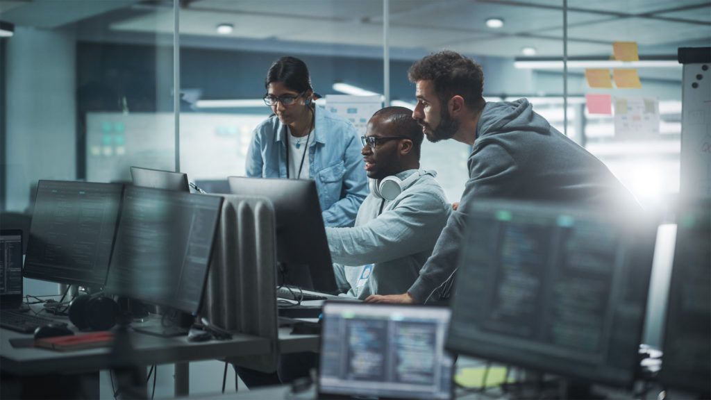 Três pessoas em um escritório olhando para um monitor de computador; um espaço de trabalho colaborativo.