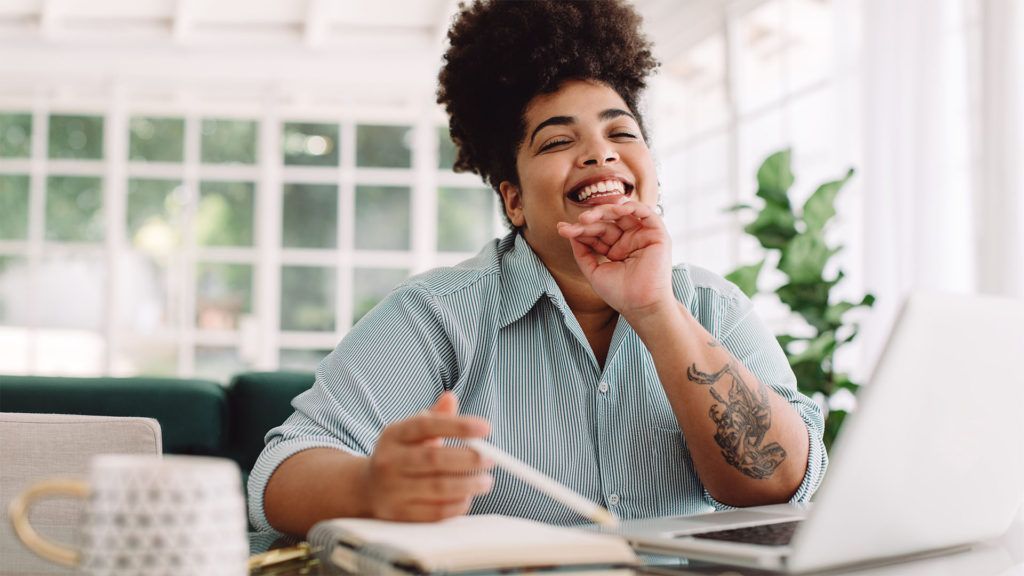 Mulher sorridente, trabalhando em um laptop em uma mesa, segurando uma caneta. Camisa listrada azul-clara, tatuagem, caderno, caneca.