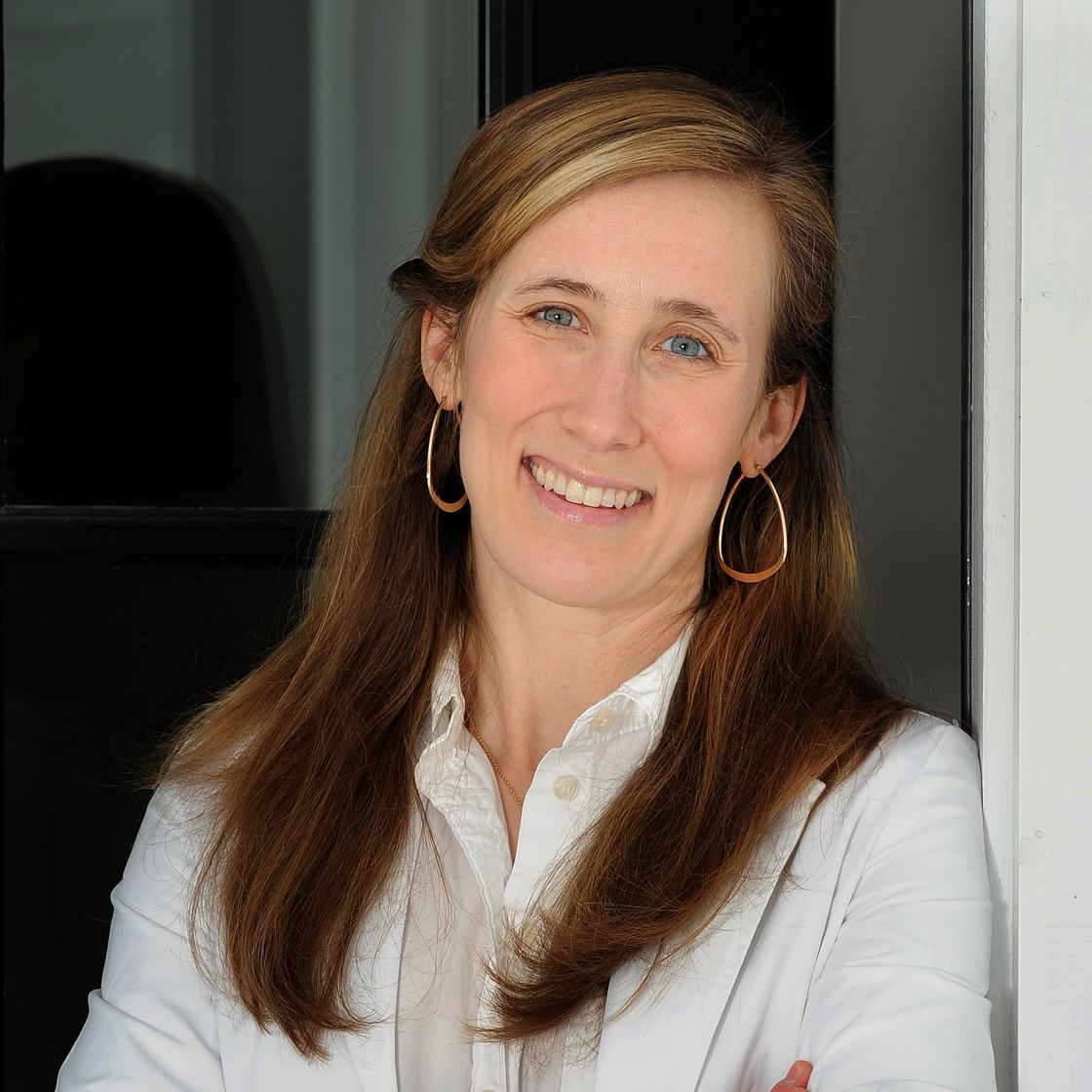 A woman wearing hoop earrings and a white jacket smiles for the camera