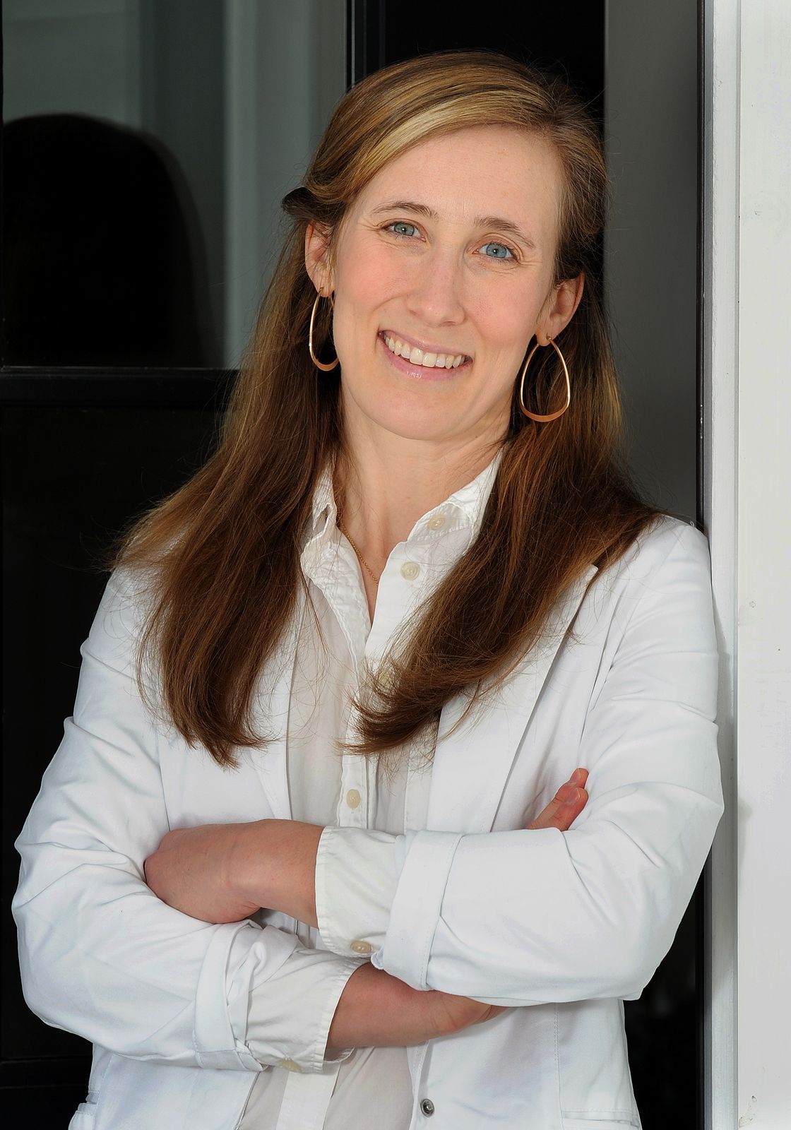 A woman in a white jacket is standing with her arms crossed and smiling.