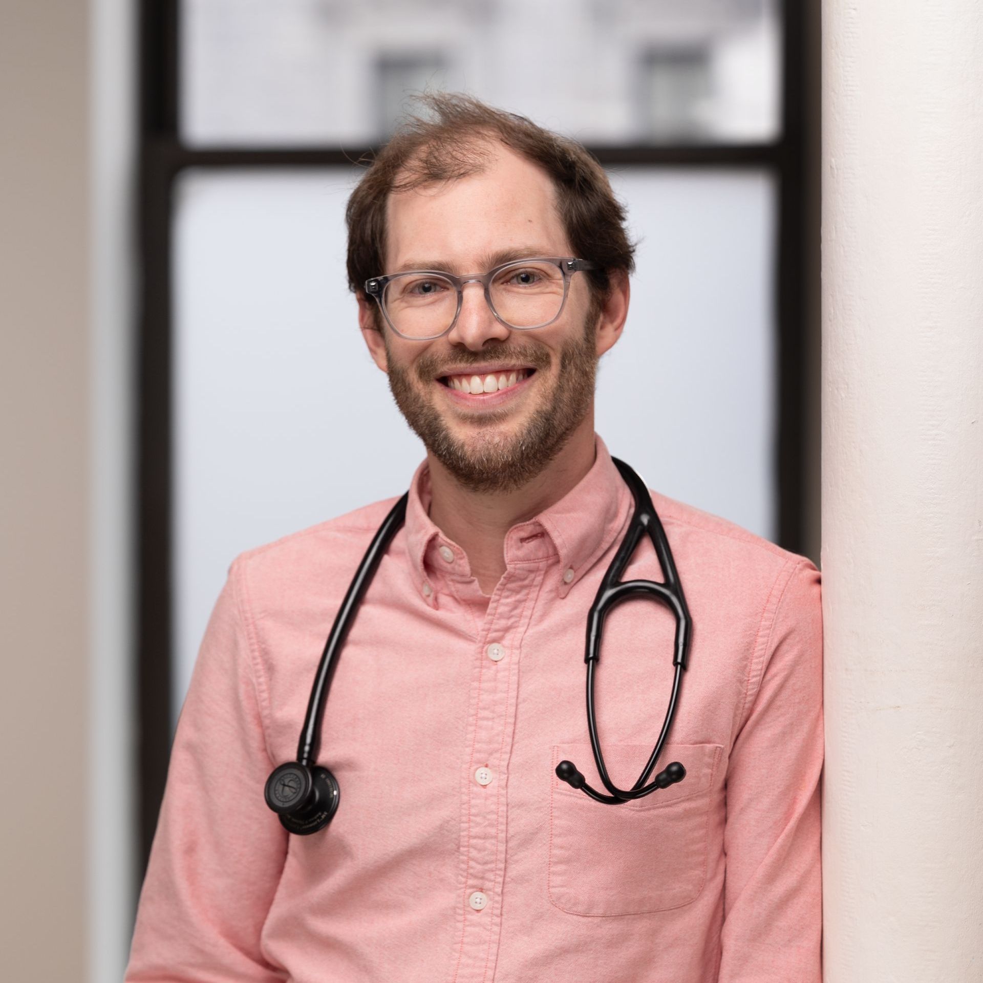 A man wearing a pink shirt and a stethoscope around his neck is smiling.