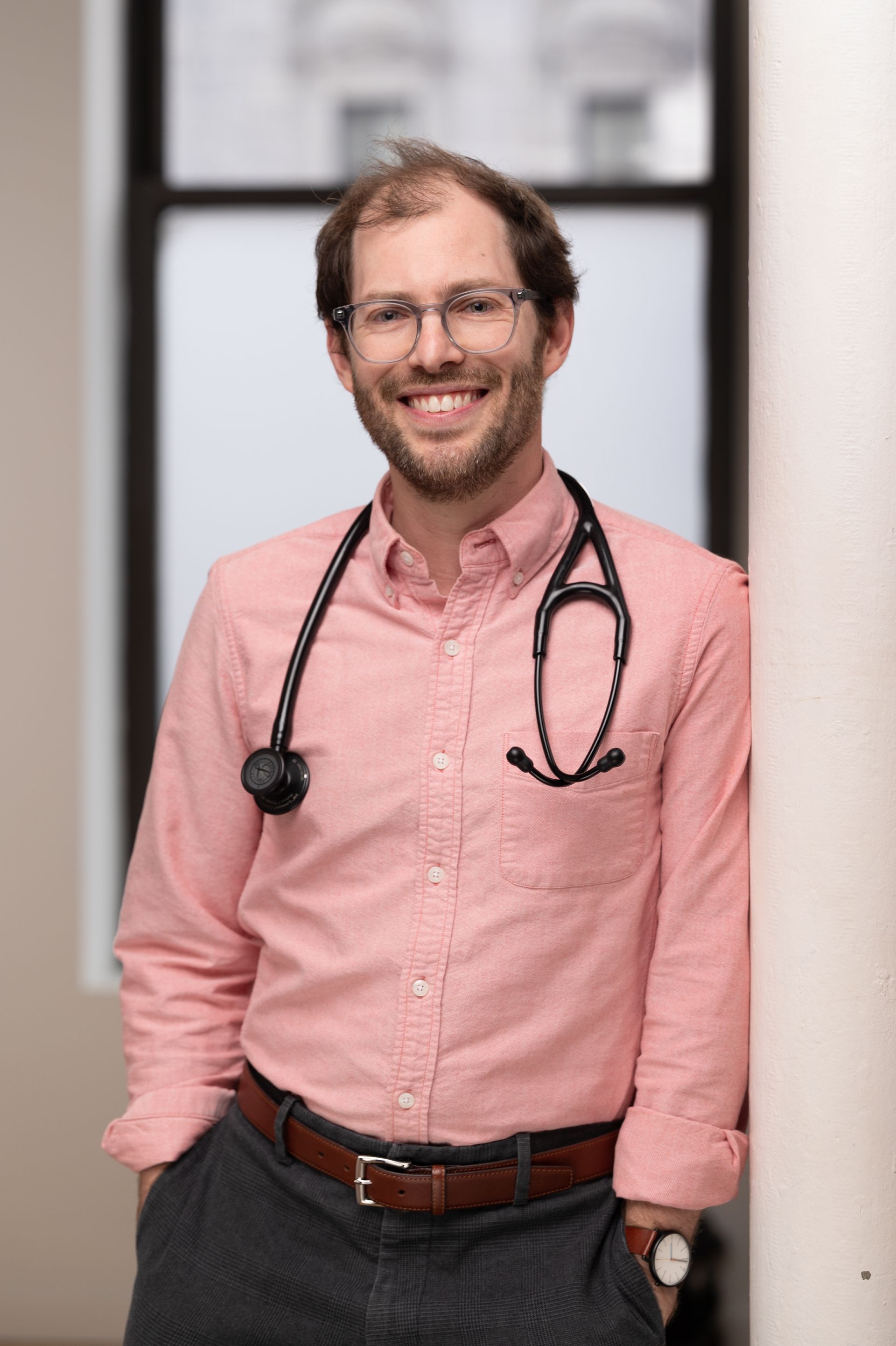A man wearing a pink shirt and a stethoscope around his neck is leaning against a wall.