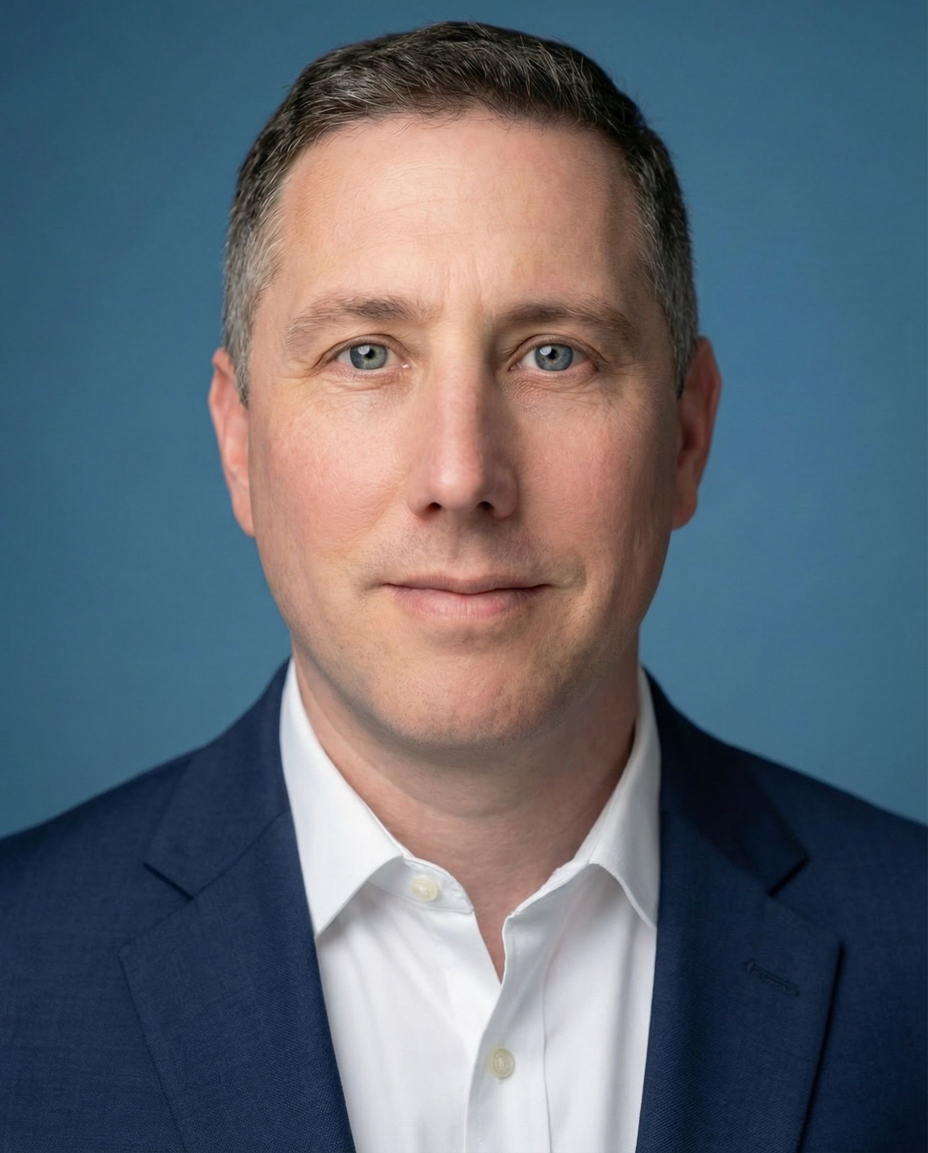 Man in a blue suit and white shirt, looking directly at the viewer, against a blue background.