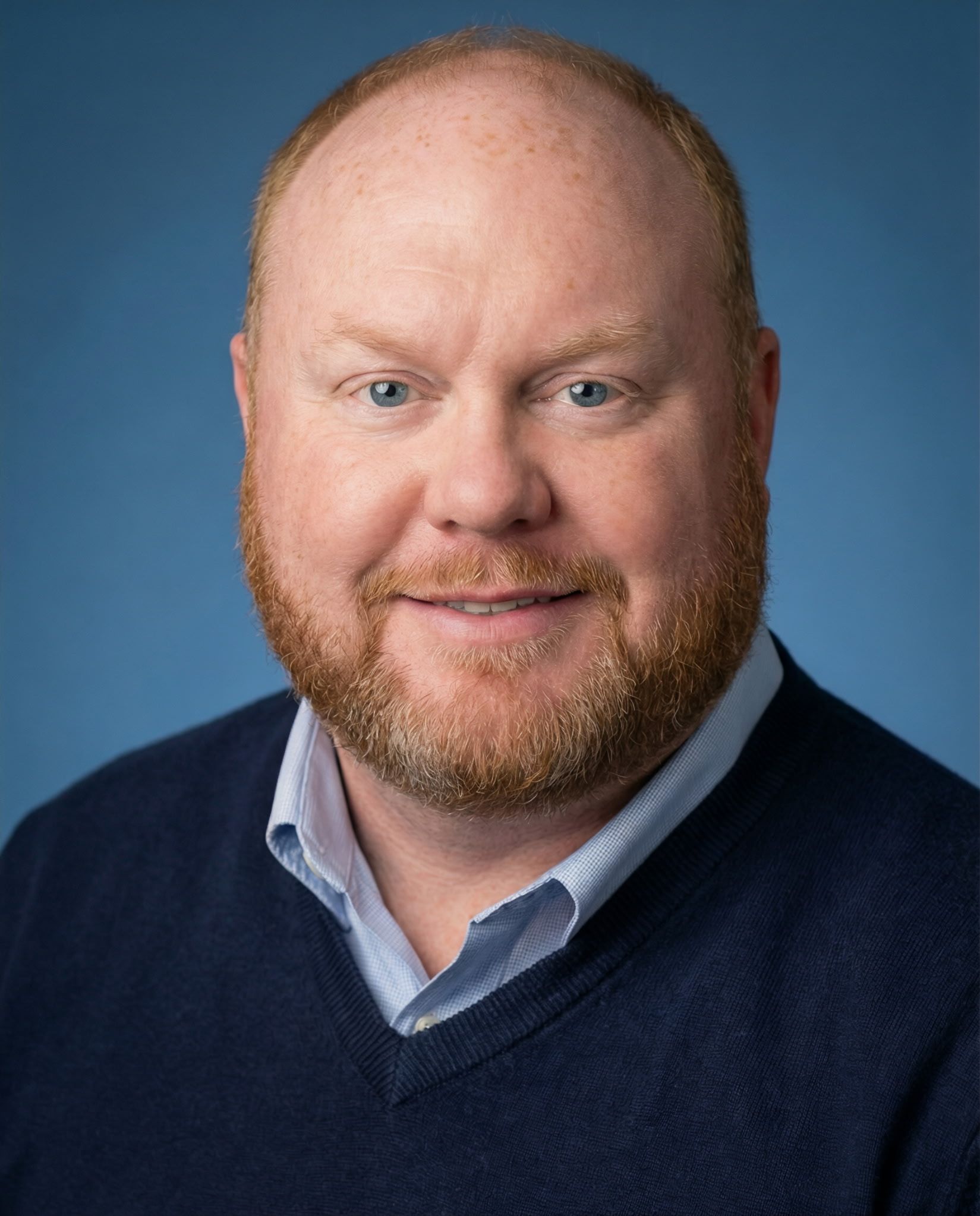 Man with a reddish beard and fair skin smiles, wearing a navy sweater over a light blue collared shirt.