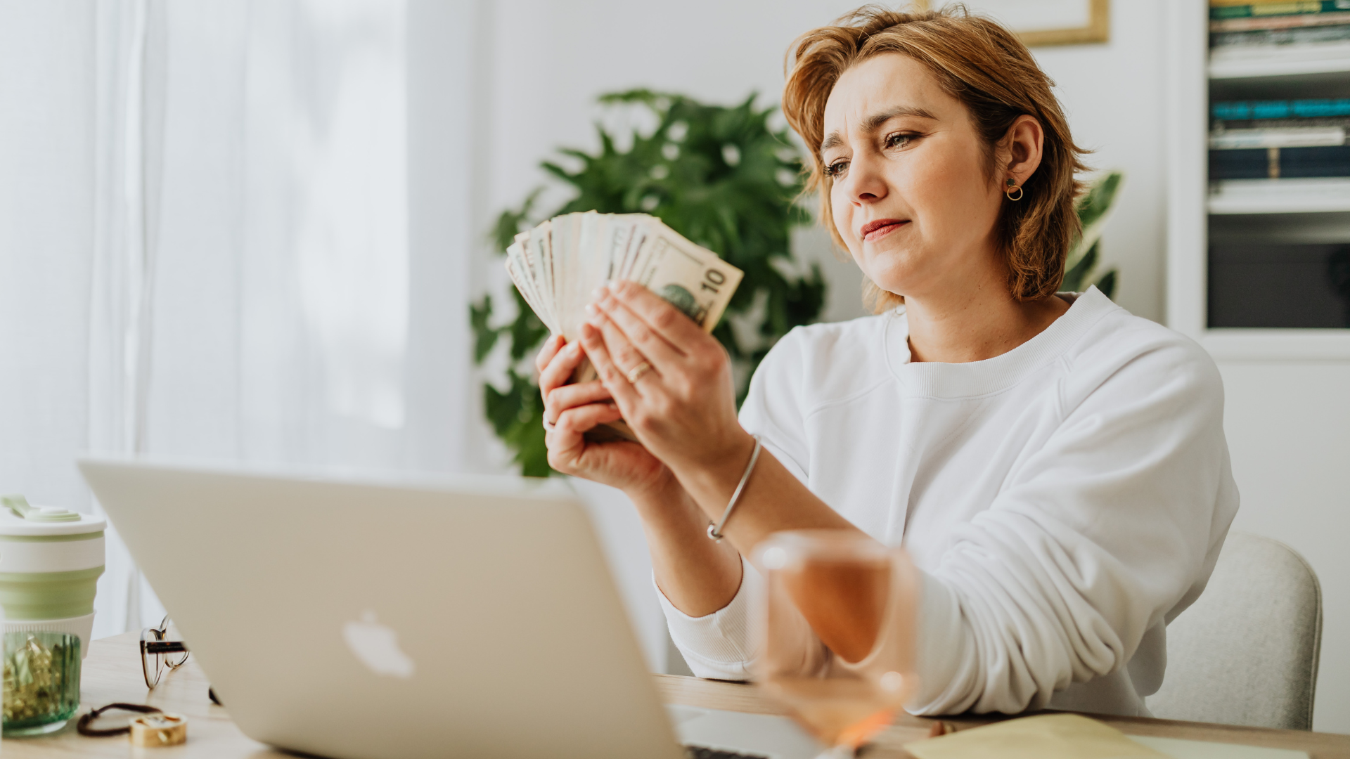 Person counting cash beside a laptop in a bright home office