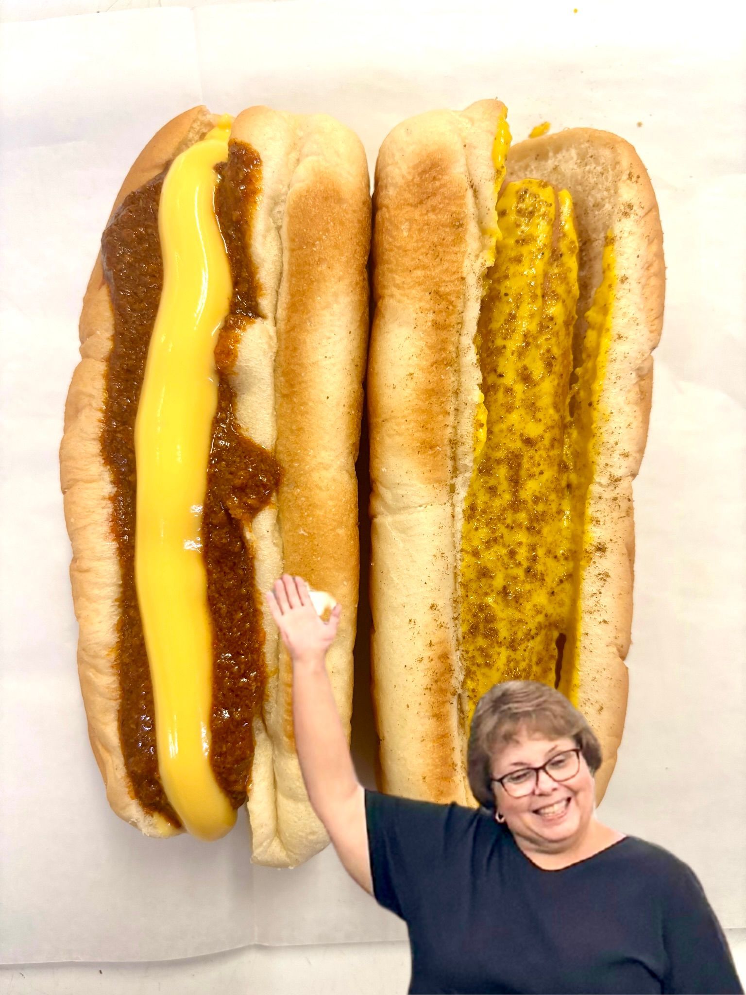 Woman smiles, pointing at two hotdogs: one with chili and cheese, one with mustard.