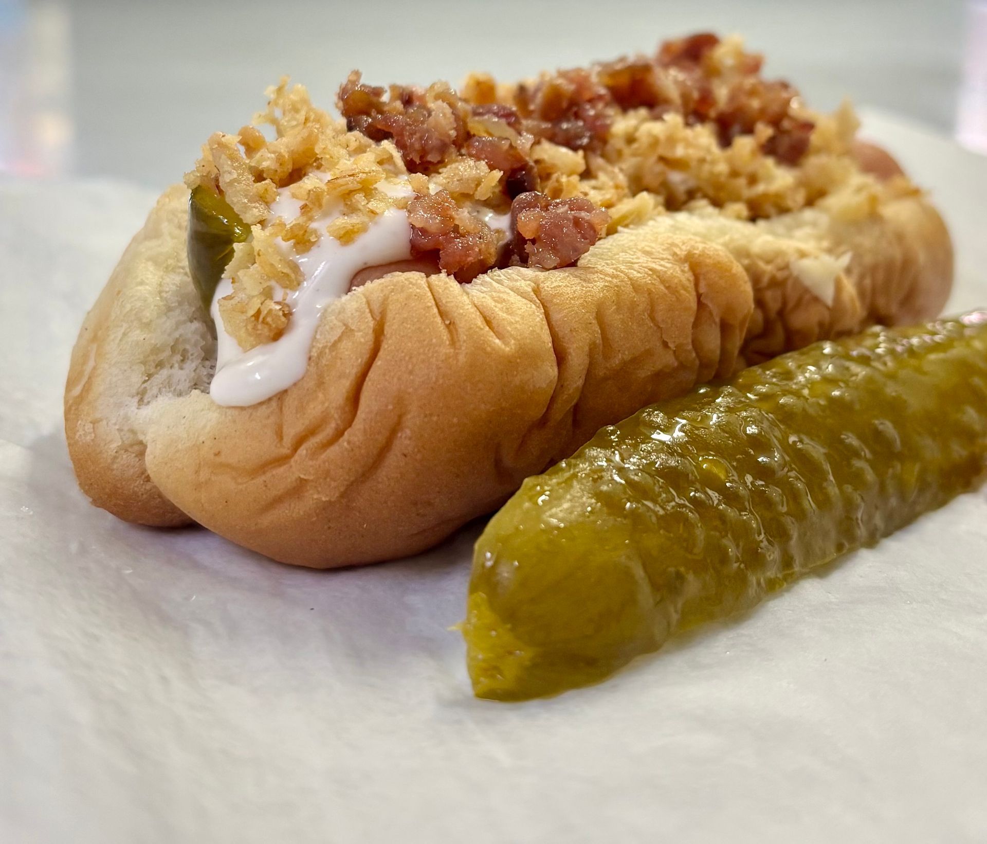 Hot dog on a bun with toppings and a pickle spear on white paper.