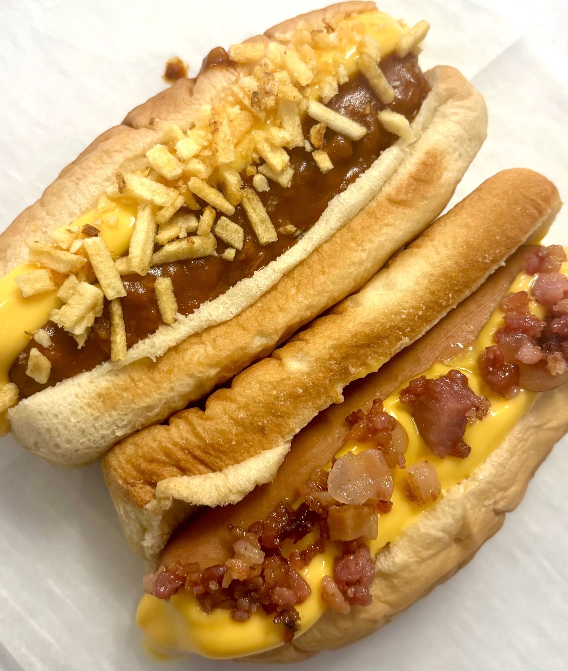 Two hot dogs: one with chili and crispy fries, the other with cheese and bacon.