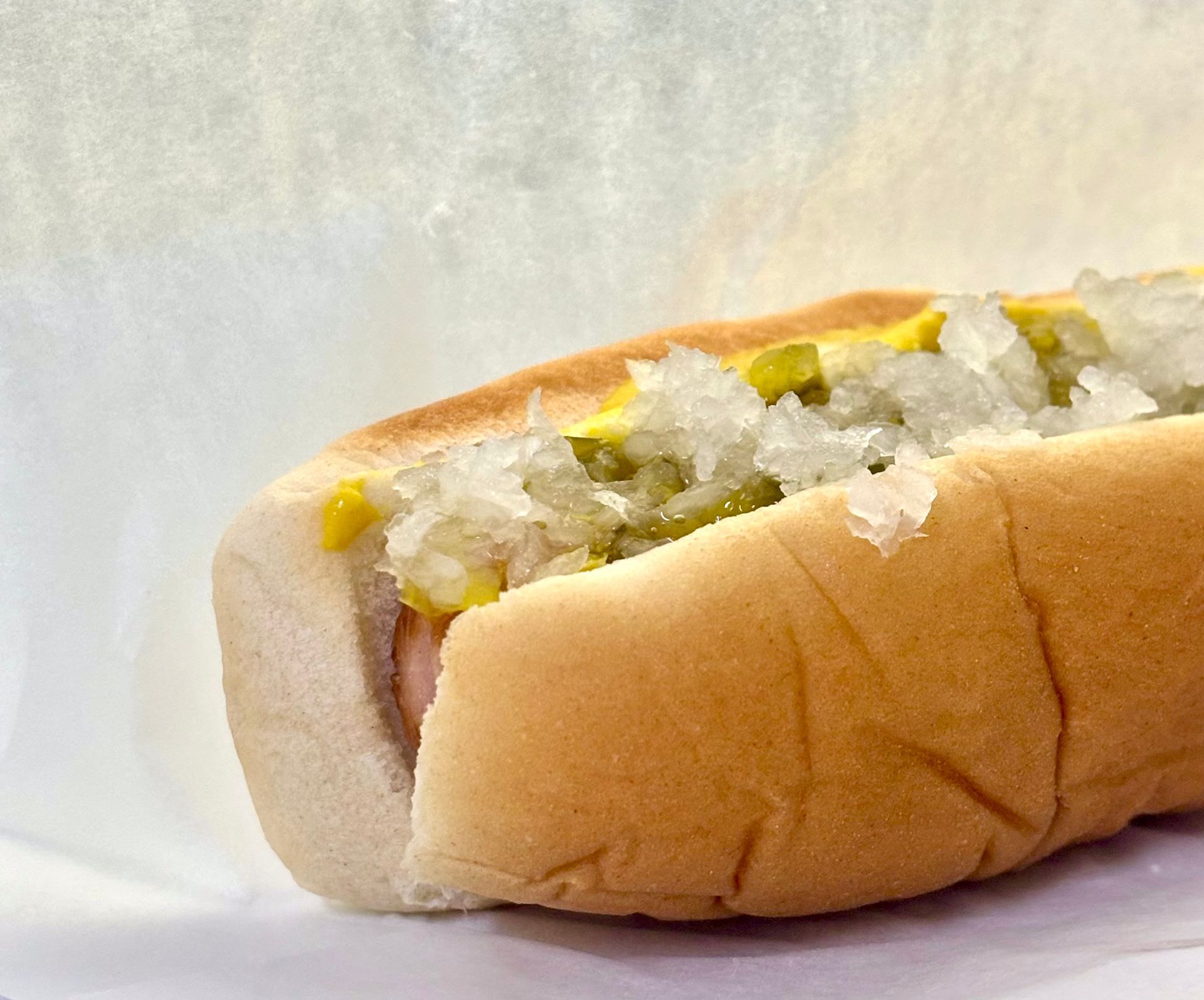 Hot dog in a bun, topped with chopped onions and relish, on white paper.