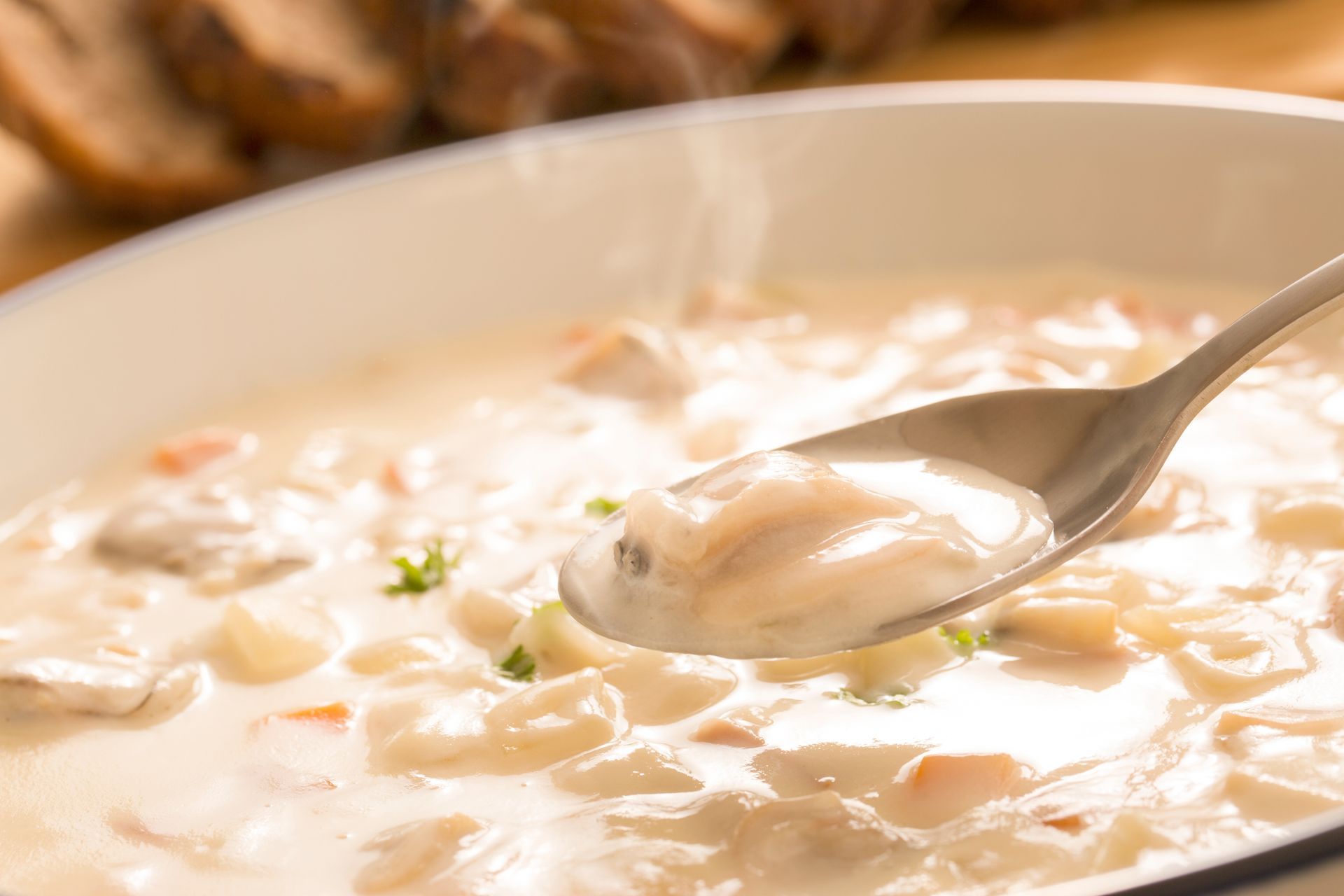Creamy seafood chowder in a white bowl, spoon lifted, steam rising, bread slices in the background.
