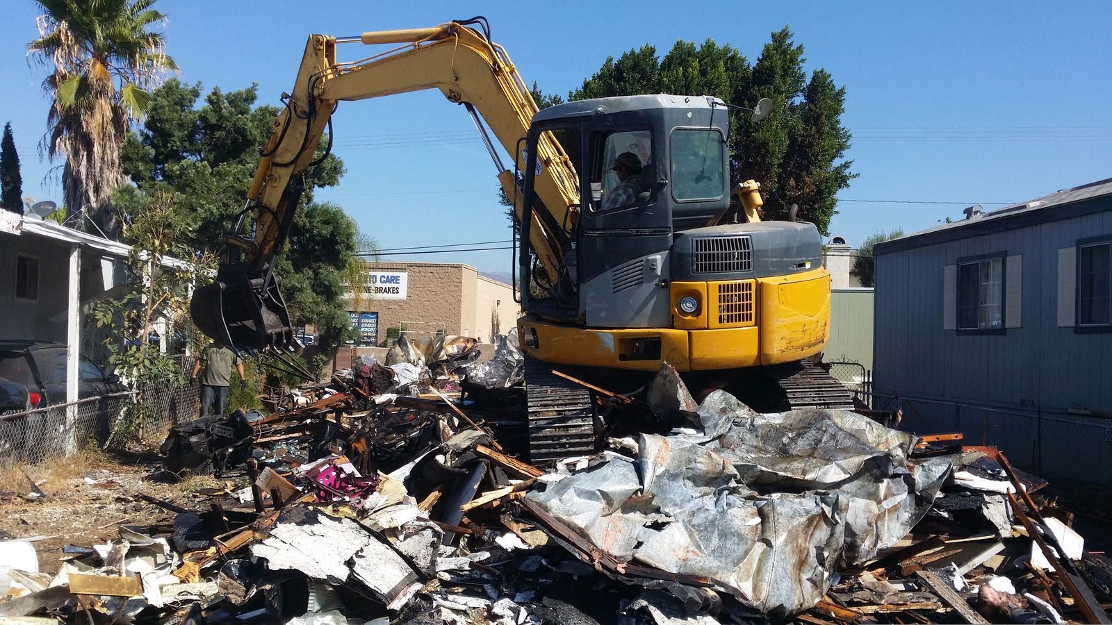 Yellow excavator demolishing rubble of a burnt structure in a sunny outdoor setting.
