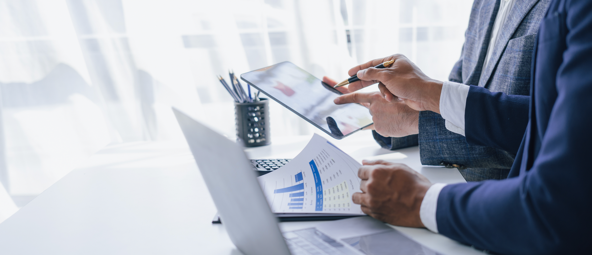 Two business people are sitting at a table looking at a tablet and a laptop.
