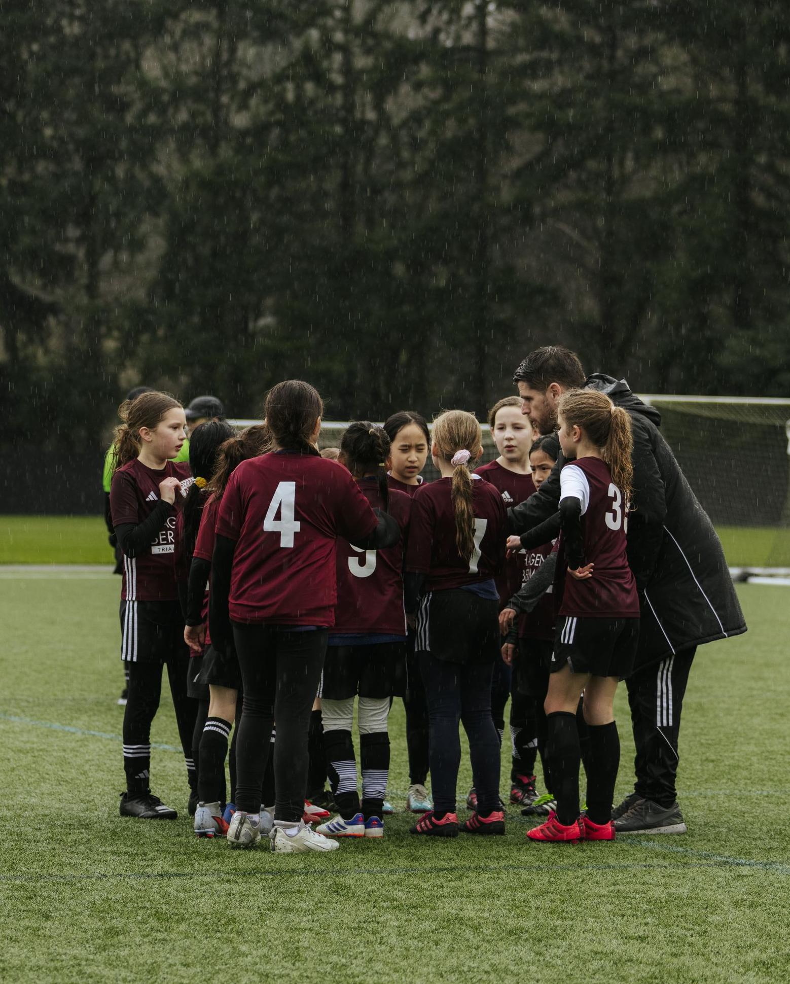 Youth soccer team huddled on a rainy field, wearing maroon and black uniforms during a break.