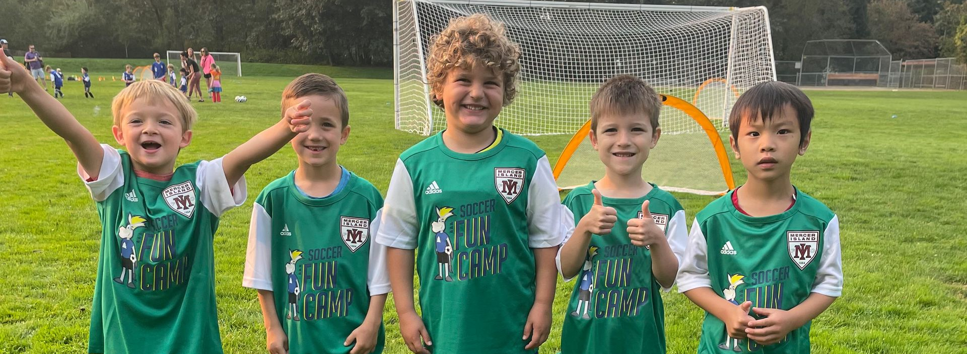 Five children in green soccer jerseys posing on a field in front of a goal, smiling and cheering