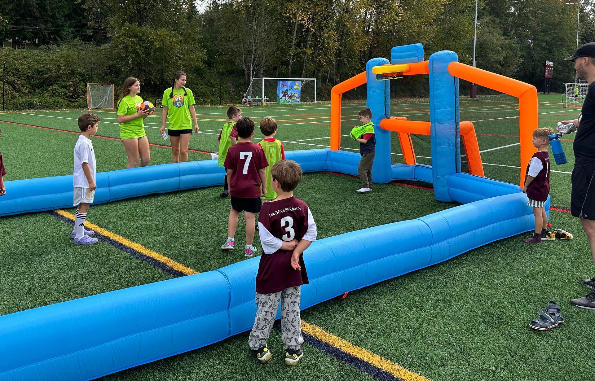 People playing inflatable outdoor sports game on a grassy field, with blue and orange barriers and goals