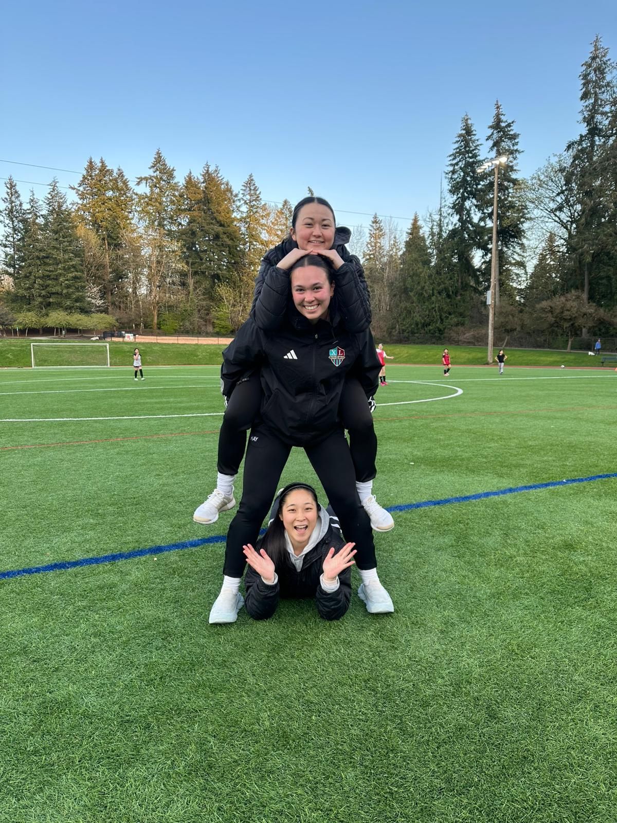 Soccer players stacked in a playful human pyramid on a green soccer field with trees in the background