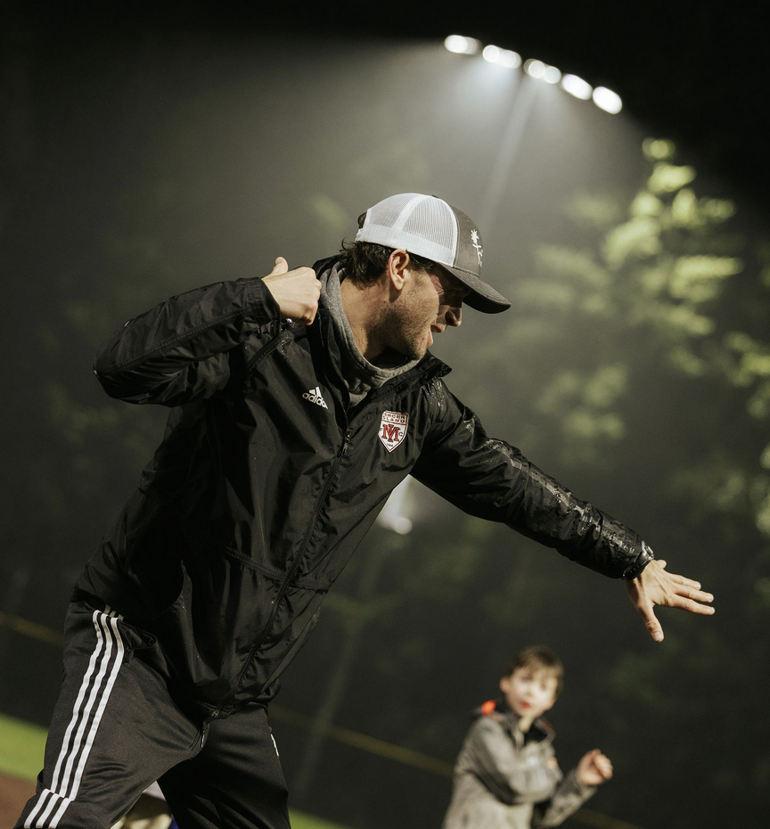 Man in black sportswear and cap dancing under stadium lights, with a blurred figure in the background