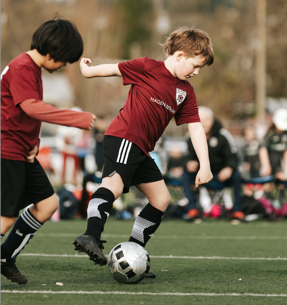 Two soccer players in maroon jerseys battle for the ball on a grass field.