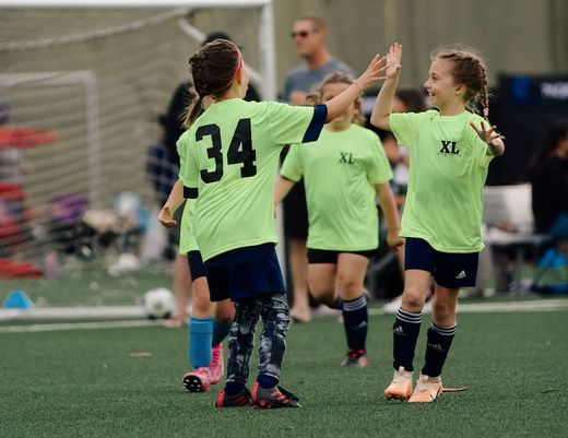 Youth soccer players in neon green jerseys celebrate with a high-five on a field.