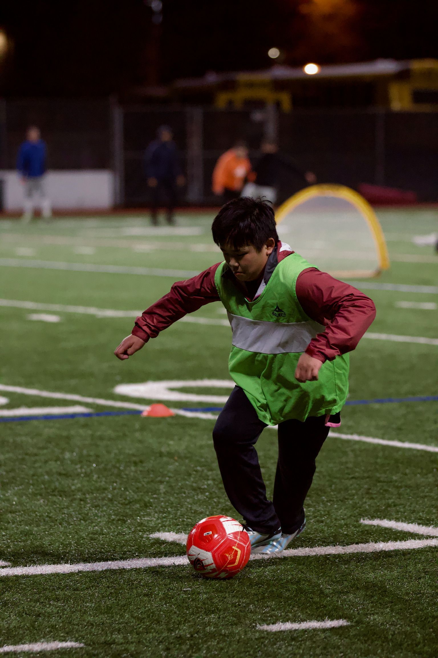 Child in green vest dribbling a red soccer ball on a lit field at night