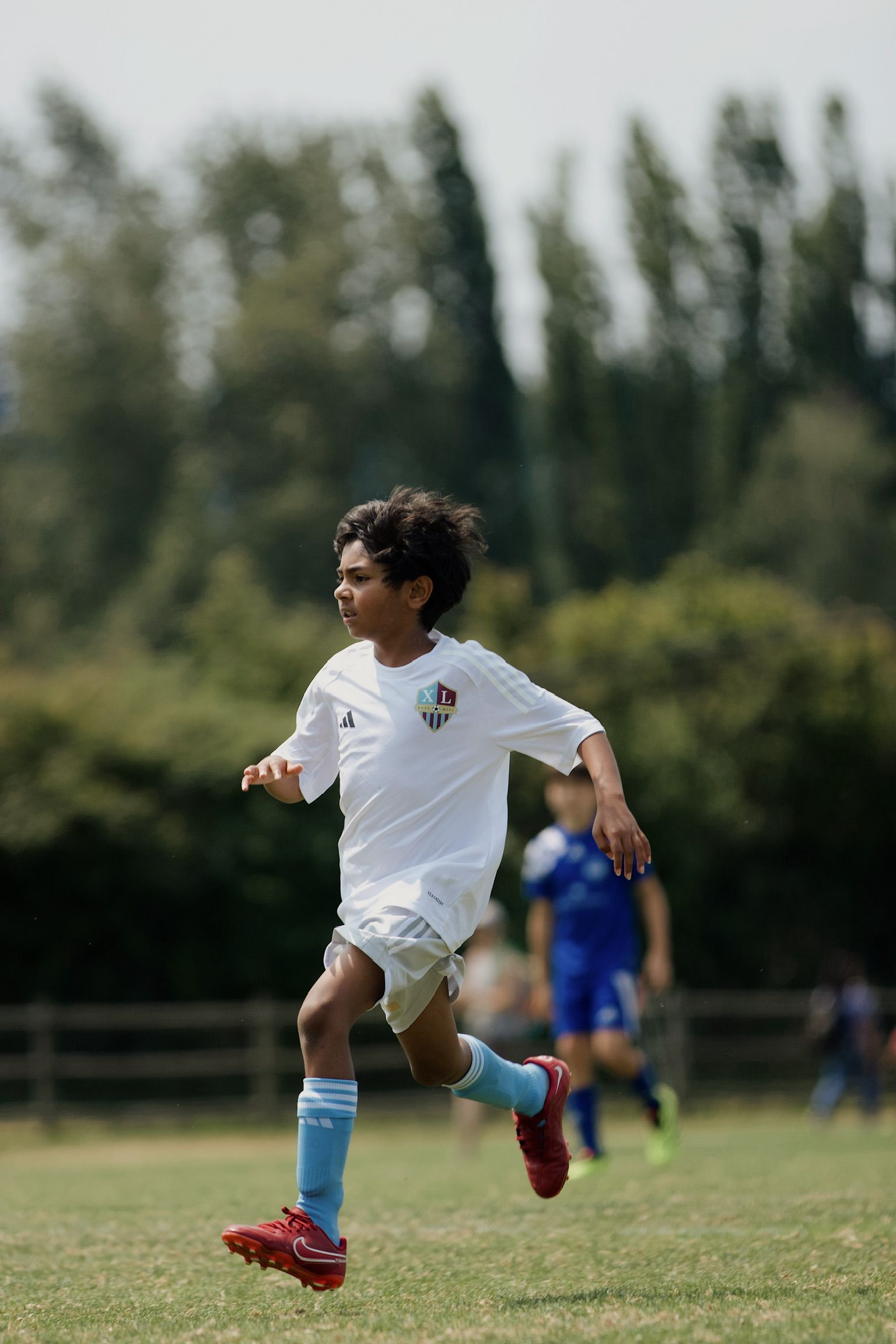 Soccer player in white sprinting on a grassy field, with blurred players and trees in the background
