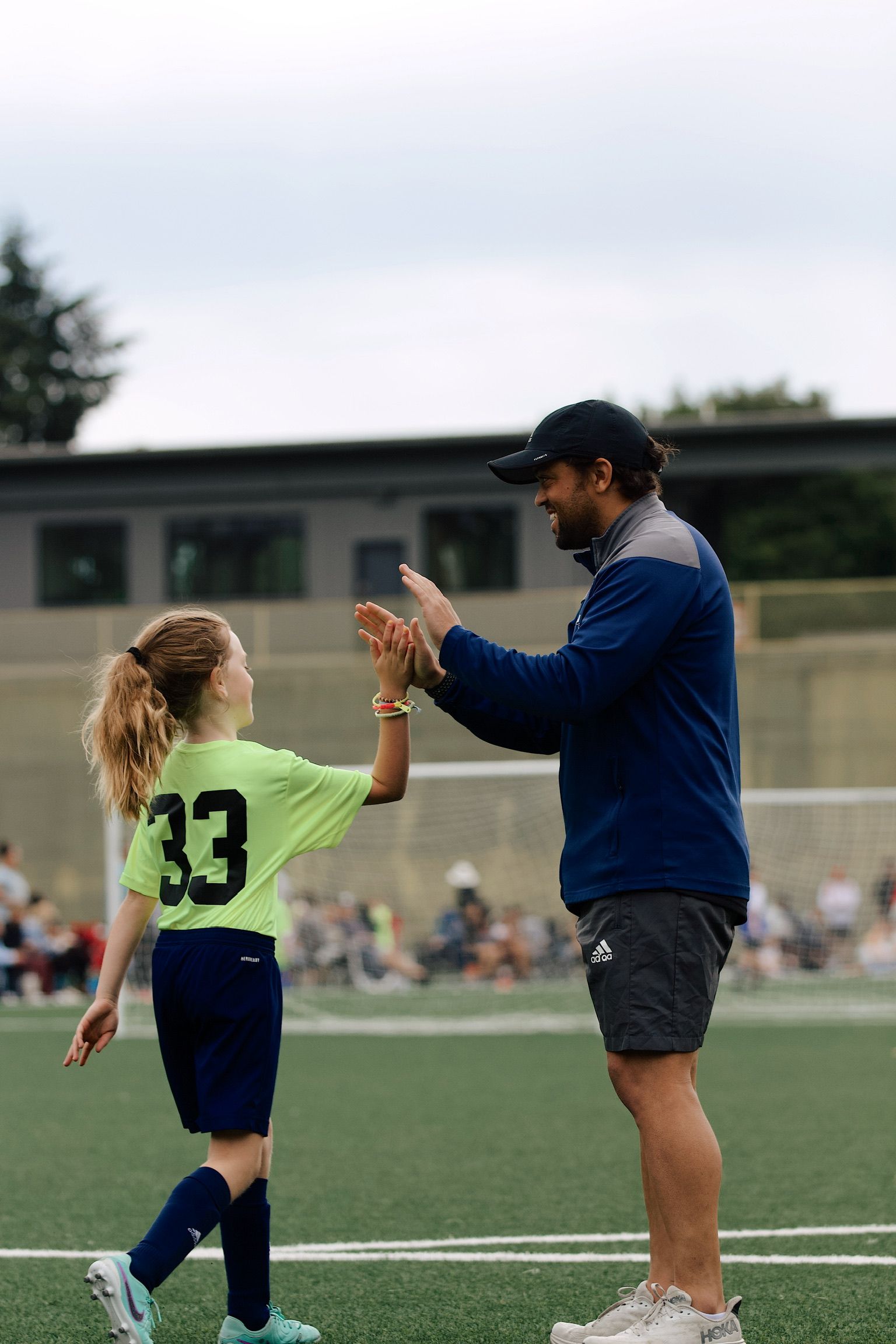 Soccer coach high-fives a young player on the field during a game, with spectators in the background