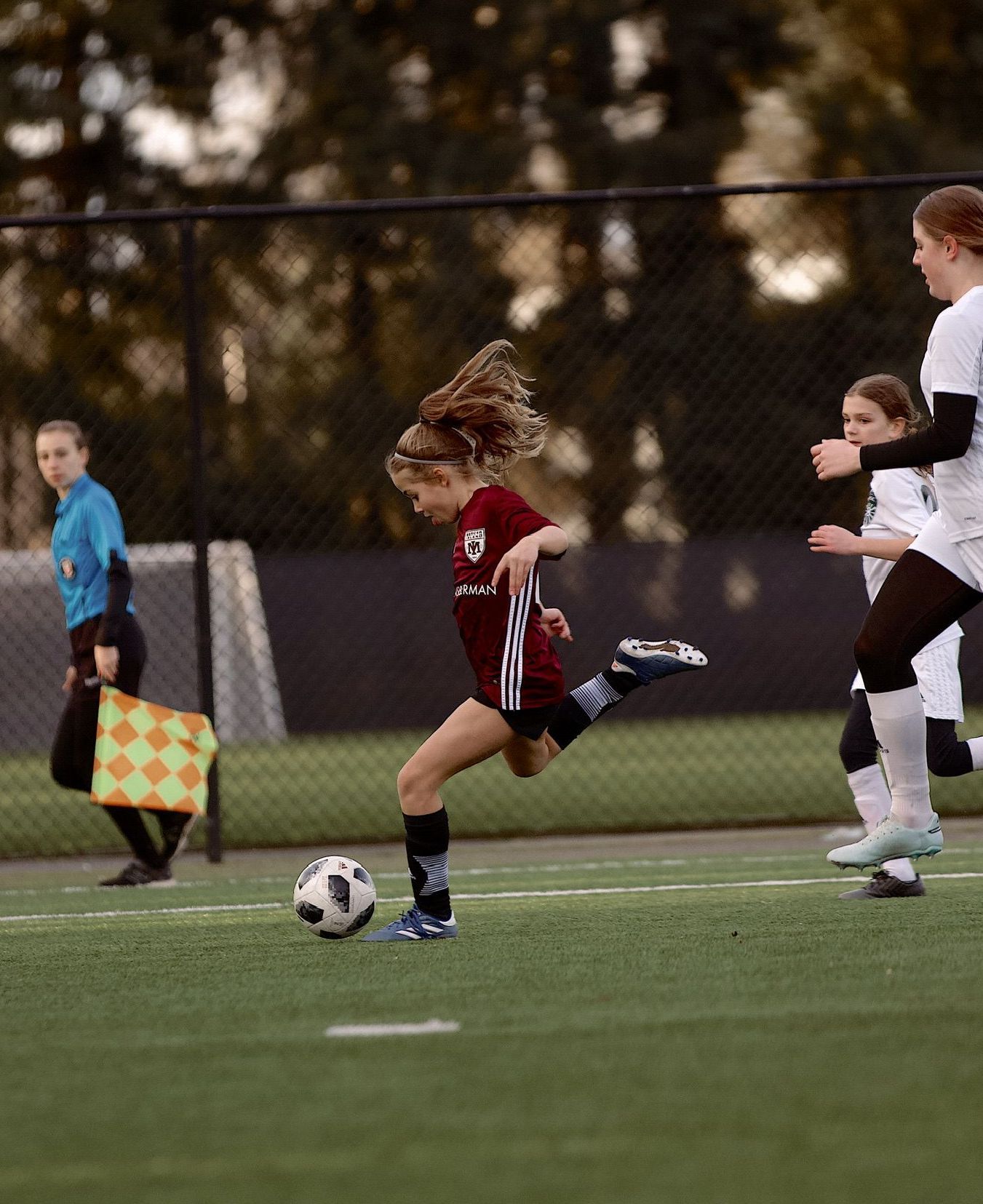 Youth soccer player in red dribbles the ball on a field while teammates and a referee watch nearby.