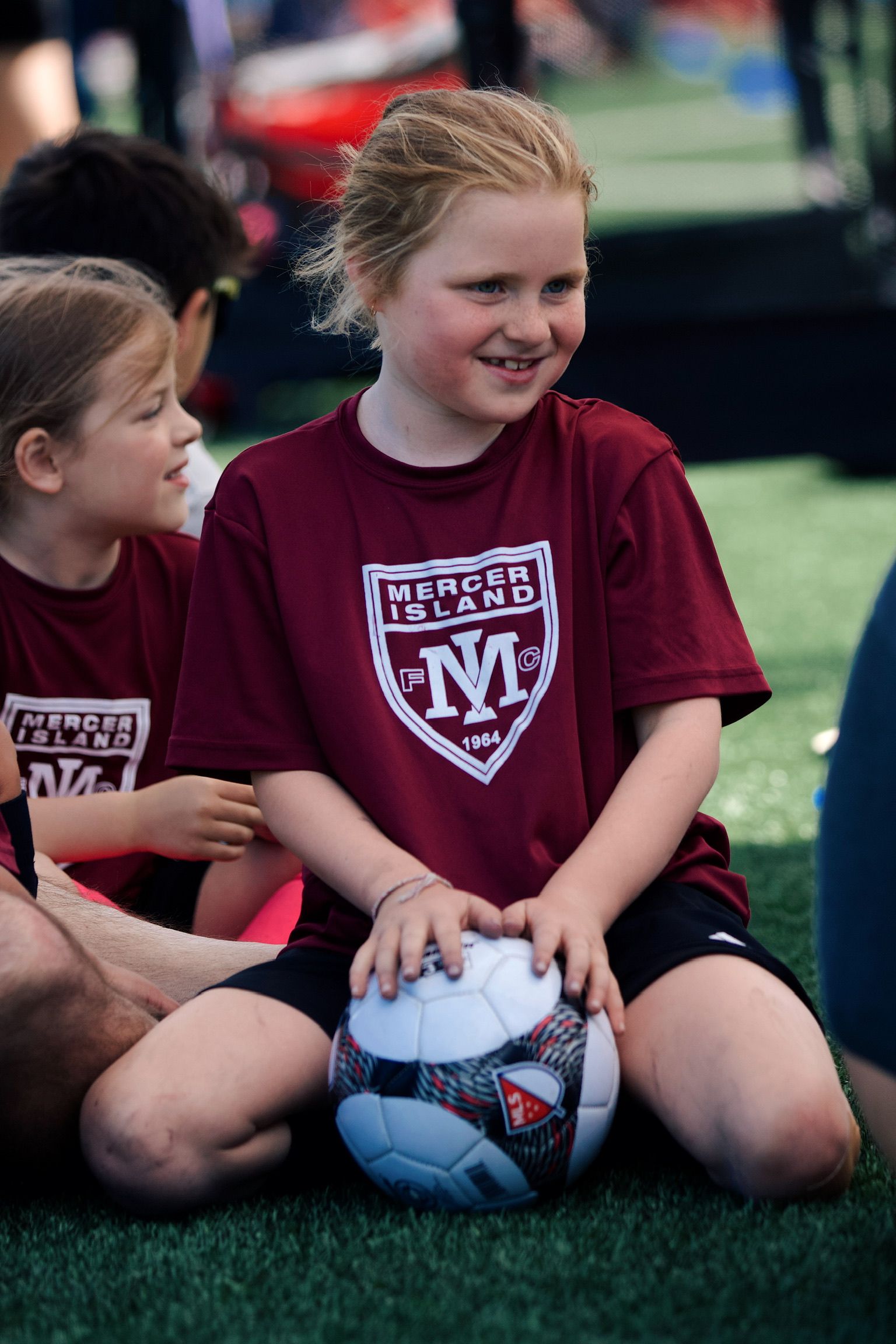 Two children in maroon sports jerseys sitting on grass with a soccer ball, one smiling