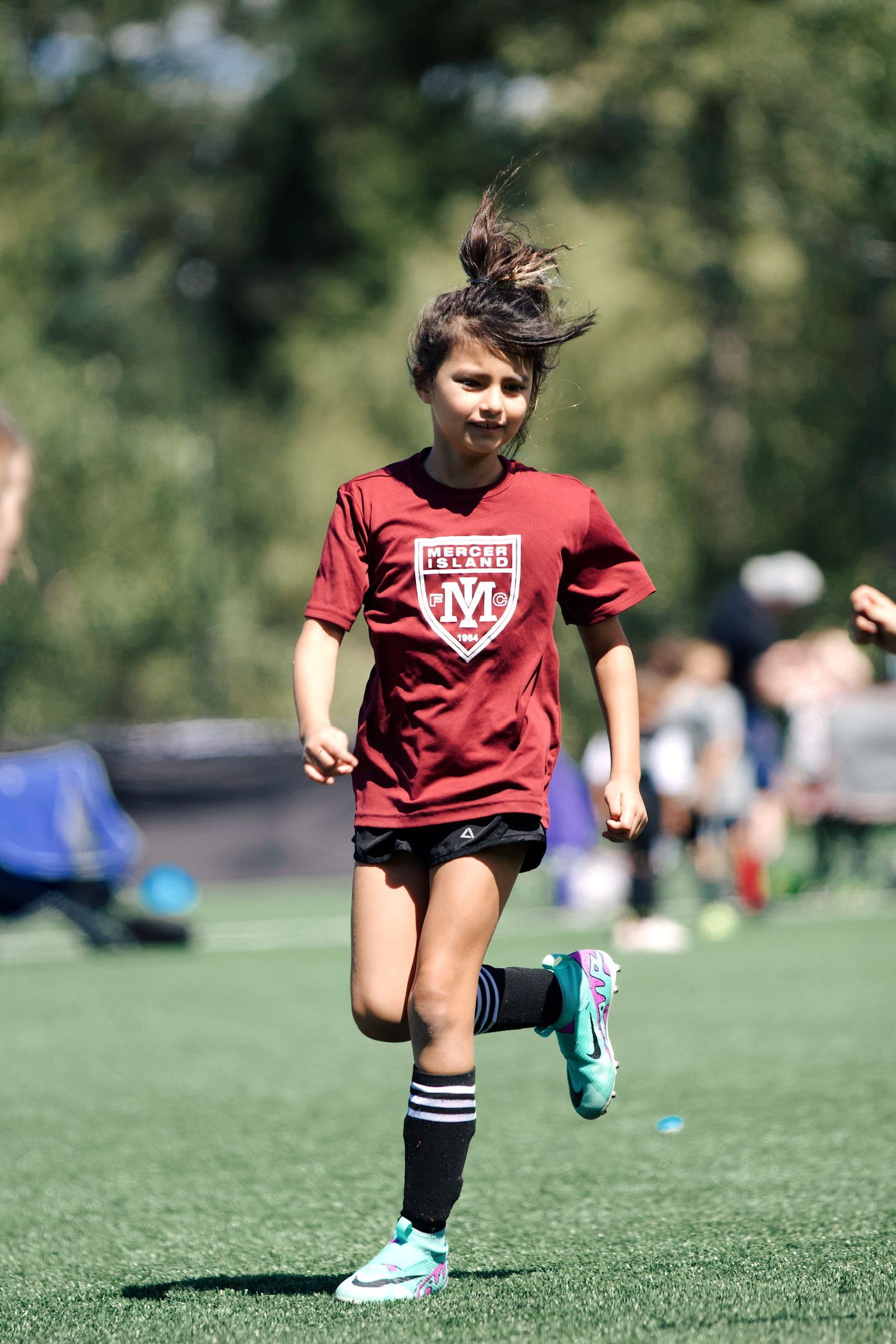 Child jogging on a soccer field in a red shirt and black shorts, with a blurred crowd in the background