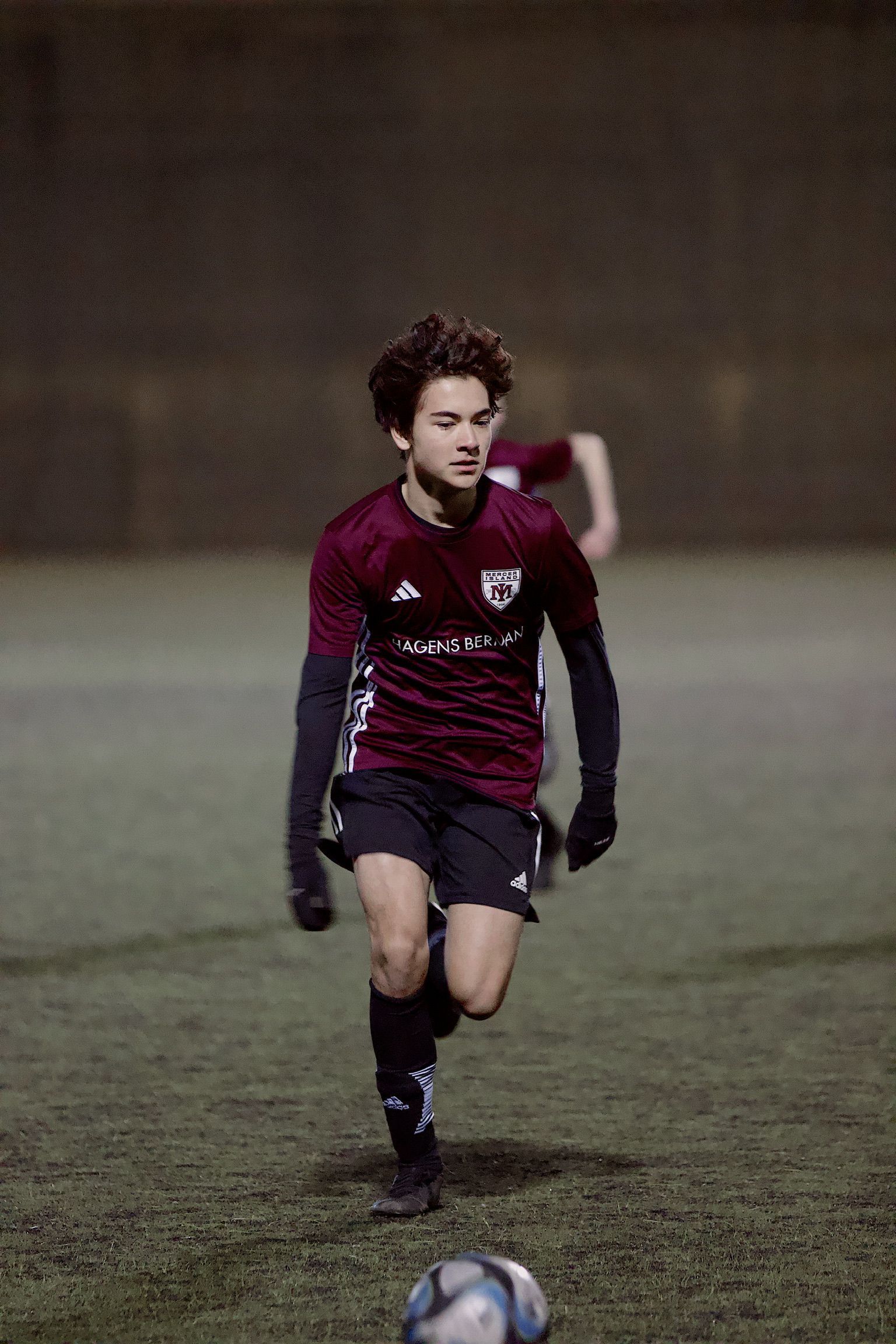 Soccer player in maroon and black uniform running toward the ball on a dimly lit field