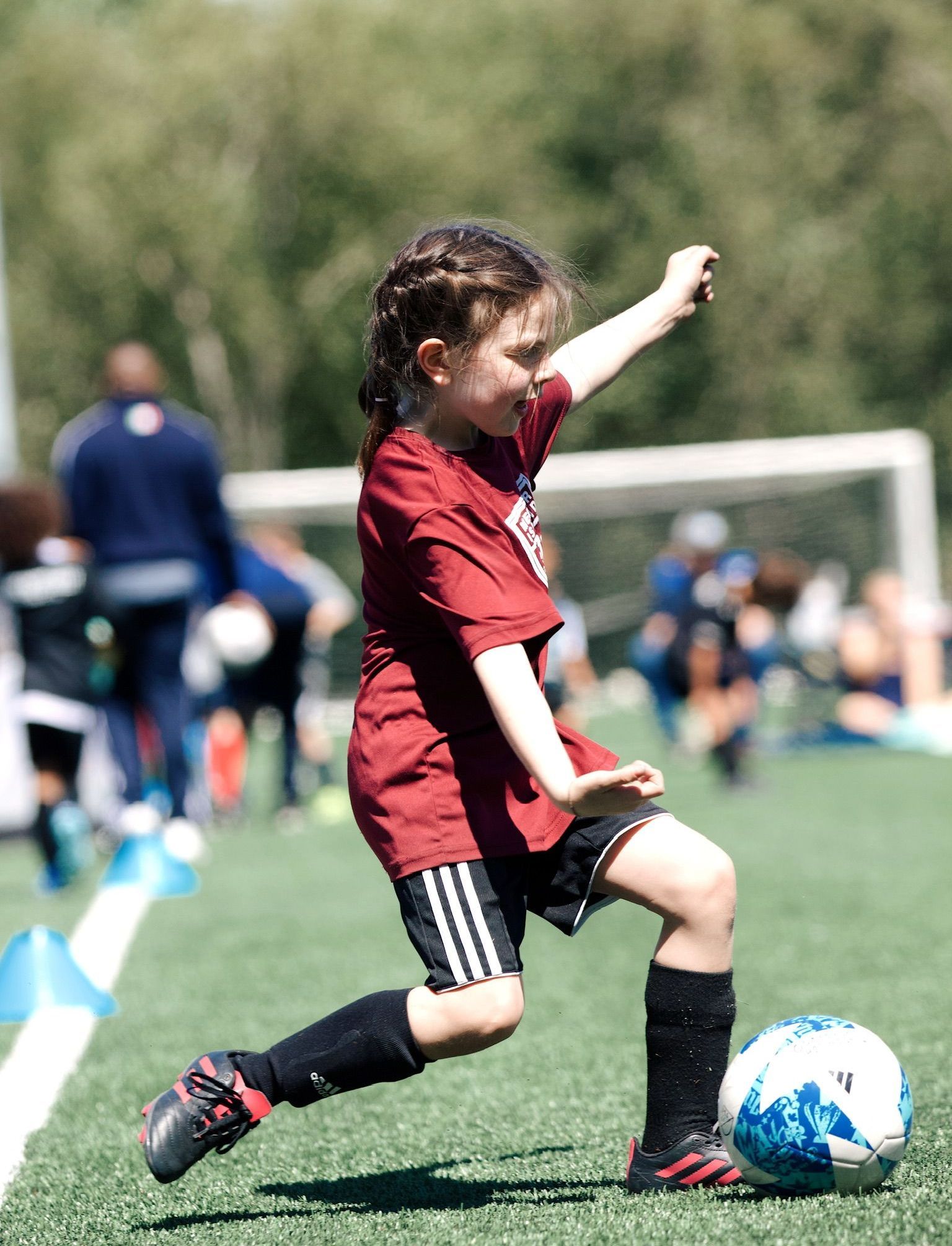 Youth soccer player in red jersey kicks a blue ball on a field with goal in the background