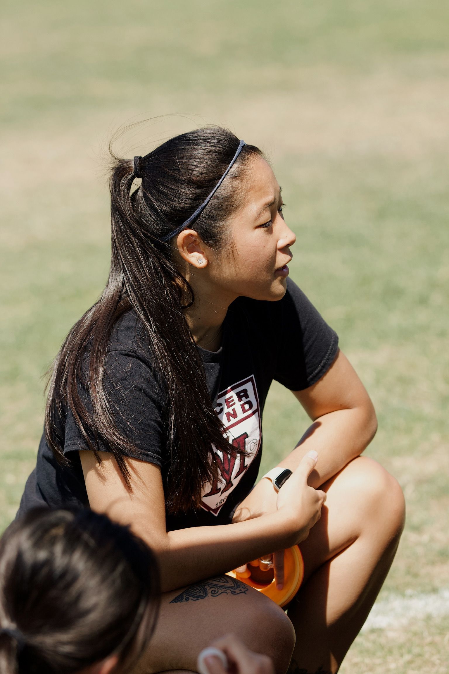Person crouching outdoors on grass, looking to the right, with another person partially visible in the foreground
