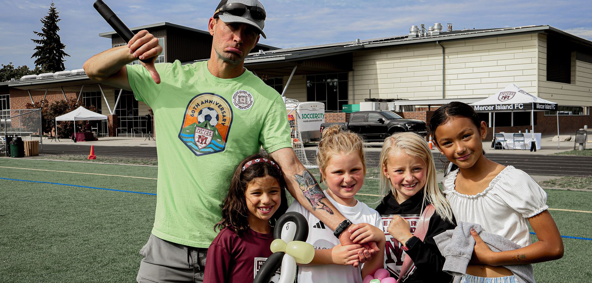 Man in green shirt with four children holding trophies on a sports field